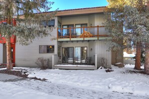 Two-story house with a balcony and sliding glass door, surrounded by snow-covered ground and pine trees. Firewood is neatly stacked on the balcony. The house has a light gray exterior.