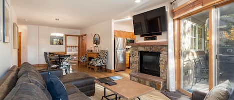 A living room with a sectional sofa, coffee table, and fireplace with a mounted TV. An open kitchen and dining area are in the background, and sliding glass doors open to an outdoor patio.