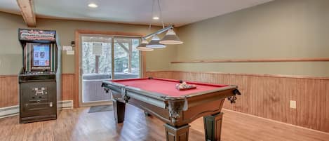 The game room, with a pool table in the center of the room, and an arcade cabinet near the back sliding glass door.