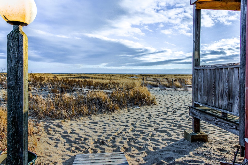 A beach view with beach grass and weathered wooden patios and lamp posts present on a calm day with blue skies and white clouds.