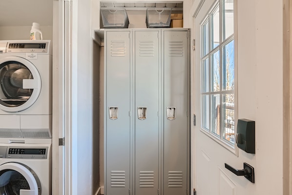 A laundry room with a stacked washer and dryer on the left, and three gray lockers next to a door with a window on the right.