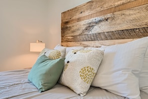 A cozy bed with white sheets and decorative pillows, featuring a green cushion and two patterned pillows, is set against a rustic wooden headboard. A bedside lamp is turned on.