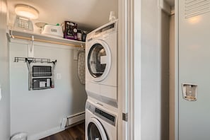 A compact laundry closet with a stacked washer and dryer, a metal shelf holding detergent and supplies, and a wall-mounted drying rack.