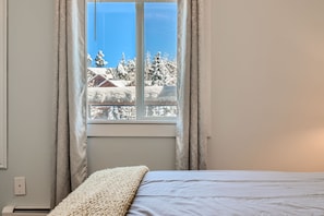 A bedroom with a neatly made bed, gray bedding, and a cream-colored throw at the end. A window with silver curtains showcases a snowy landscape outside.