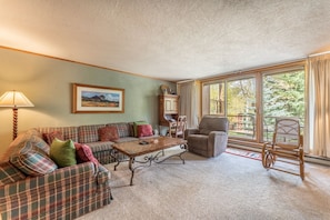 Spacious living area with cozy furniture, wood-burning fireplace, flat screen TV and balcony access.