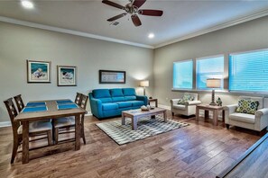 Large great room with wood tile floors, sofa, two side chairs, occasional tables and lamps and dining table for 4