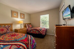 Bright bedroom featuring two beds with colorful patterned bedding, a dresser, and a window providing natural light.
