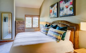 Bedroom with white cover and green and patterned decorative pillows.  Tall, wood chest of drawers next to window in background.