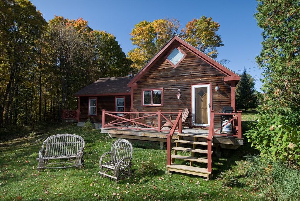 A quaint log cabin in Stowe for rent has a front porch area with additional seating in the yard in front