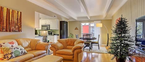 A living room with beige walls features a tan leather sofa set, a coffee table, a flat-screen TV, a small Christmas tree, and an adjacent dining area with a round table and chairs.