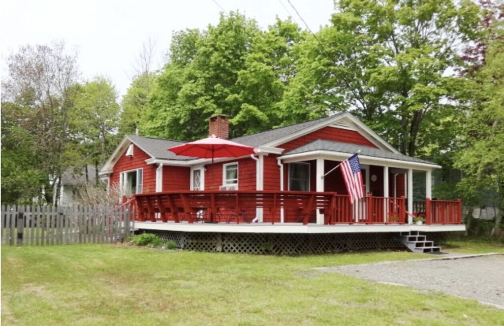 A bright red vacation rental in Bar Harbor, Maine has a wrap around porch, American flag flying, and is surrounded by green grass and trees.