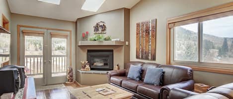 A cozy living room features a leather sofa, armchair, wooden coffee table, and TV. Skylights and a large window illuminate the space, which includes a fireplace and decor of forest-themed art and figures.