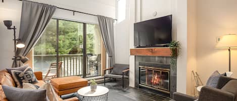 Living room w/ wood-burning fireplace and cable TV leading out to balcony w/ small Webber BBQ
