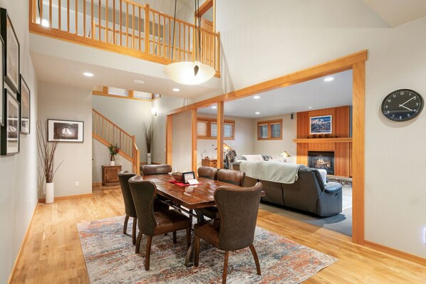 This open-concept space features a wooden dining table for six, a cozy living room with a plush sectional, and a warm fireplace framed by natural wood accents. The high ceilings and soft lighting create an inviting and relaxing atmosphere.