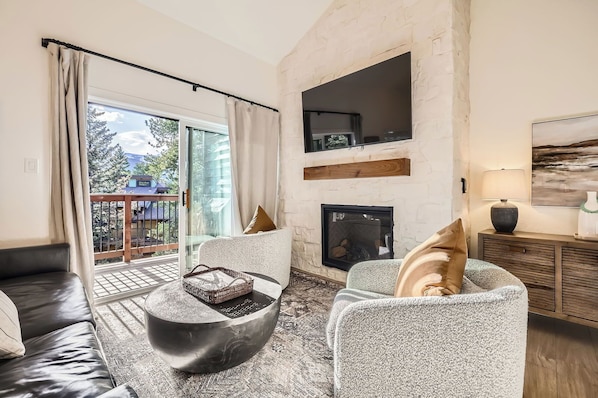 Cozy up in the stylish living room, featuring modern furnishings, a unique antler chandelier, and floor-to-ceiling windows that open to a private balcony with breathtaking forest and mountain views.