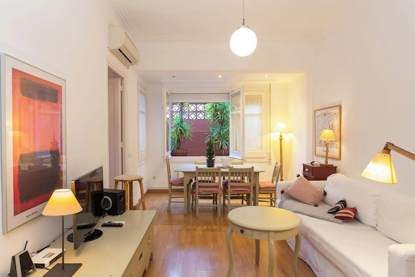 This bright and airy living room features a large window that provides natural light and a view of a charming courtyard. The room is furnished with a comfortable white sofa, a coffee table, and a dining table with chairs for 5 people. It is perfect for re
