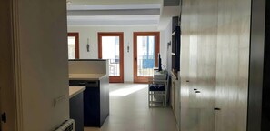 A view from the hallway of the apartment, showcasing the open plan kitchen and living area. The kitchen features a sleek and modern design with dark blue cabinetry and a white countertop. Two sets of French doors lead to a balcony, inviting natural light 