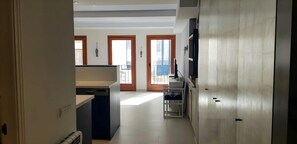 A view from the hallway of the apartment, showcasing the open plan kitchen and living area. The kitchen features a sleek and modern design with dark blue cabinetry and a white countertop. Two sets of French doors lead to a balcony, inviting natural light 