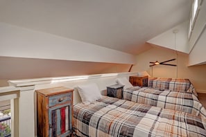A loft bedroom with two single beds, plaid bedspreads, and two rustic nightstands under a sloped ceiling. A ceiling fan is visible above the beds.