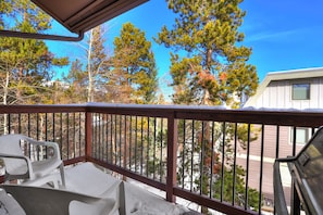A snowy balcony with two white chairs, a wooden railing, and a clear view of nearby trees and buildings under a bright blue sky.
