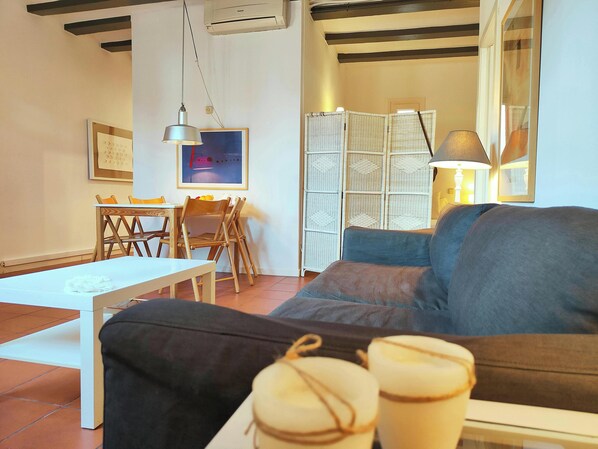 The spacious living area features exposed wooden beams, a comfortable couch, and a white coffee table. The apartment has a modern aesthetic with a touch of warmth, featuring a light-colored wicker room divider and a framed abstract painting on the wall. A