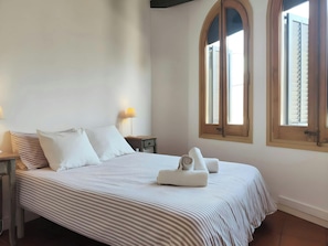 This bedroom features a comfortable queen-size bed with crisp white linens and a striped duvet cover. Natural light streams in through the two arched windows with wooden shutters, adding a touch of traditional charm to the space. The bedside table with a 
