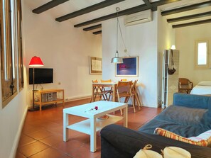 The living room features exposed wooden beams, a large window with wooden shutters, a flat screen TV, a comfortable sofa, and a dining table with four chairs. A white coffee table is placed in the center of the room, and there is a red lamp to the left of