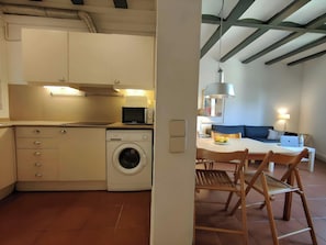 The apartment features a modern kitchen with white cabinetry, a built-in microwave, and a washing machine. The kitchen is adjacent to the dining area, which features a white table and four wooden chairs. The dining area is open to the living room, which h