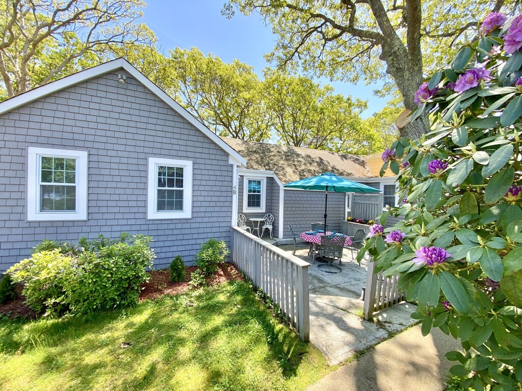 A blue VRBO in Cape Cod in summer is surrounded by trees, flowering bushes, and grass.