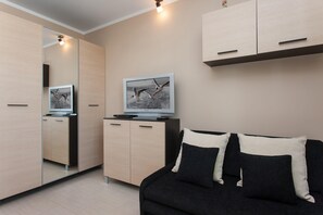 The living area from a different angle, showing a painting on the wall and a small wardrobe with light wood tones. The black couch and pillows add contrast to the light surroundings.