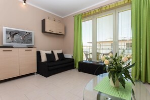 A closer view of the black couch with decorative white and black pillows, a glass coffee table, and a large window with green curtains. The room feels bright and inviting.