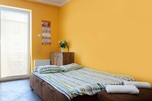 A bedroom with a single bed covered by a striped blanket, placed against a yellow wall. There’s natural light streaming in through a large window, and a plant adds a decorative touch.