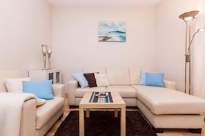 Another view of the living room featuring a light-colored sectional sofa and a blue throw. The design is simple, with soft, neutral tones.
