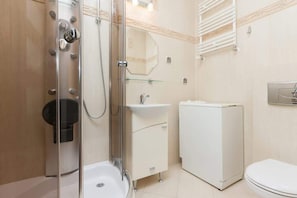 A compact bathroom with a glass-enclosed shower, toilet, and a small washing machine. The design is practical, with light-colored tiles for a clean, modern look.
