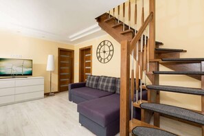 Bright, spacious living room with a chest of drawers on which stands the TV.  To the right is a corner sofa bed above which is a village clock. In the right corner there are stairs leading upstairs. 
