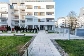 External view of the building. In front of the building you can see the well-kept lawn and paths leading to the entrance. The building is modern, with balconies and a bright facade.