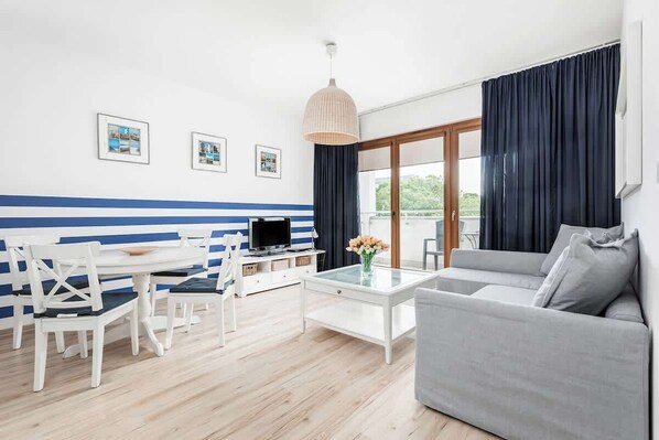 
Spacious living room with grey sofa bed. A white table with a round top is visible on the right. Four white chairs stand at the table. Pictures with a seaside landscape hang on the wall. 