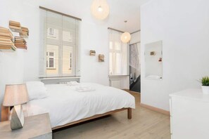  A bright bedroom with a large double bed, white linens, and a simple nightstand with a lamp. The room has large windows letting in plenty of natural light.