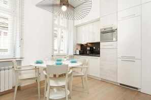 An open-plan living room and kitchen, featuring a teal sofa, dining table, and sleek white cabinets. A brick accent wall adds warmth to the space.