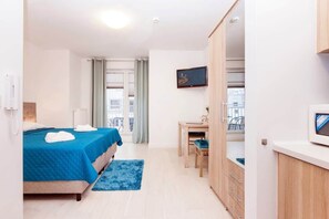 A studio-style room with a bed on the left and a kitchenette in the background. The kitchenette includes a small counter, cabinets, and basic appliances, making the space feel compact yet functional.