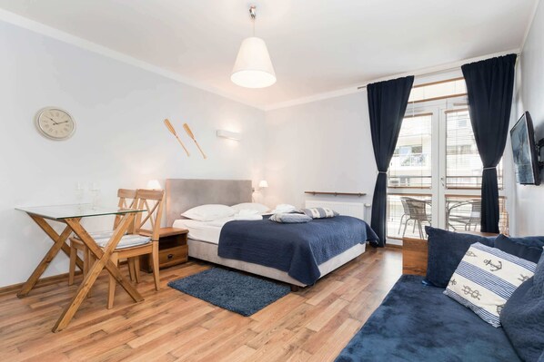 The studio features a neatly made double bed with dark blue bedding and a window with dark curtains. The room has a simple and cozy atmosphere with natural lighting.
