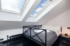 A bedroom with a double bed placed under a skylight, providing ample natural light. The minimalist decor and black bed frame give it a modern aesthetic.