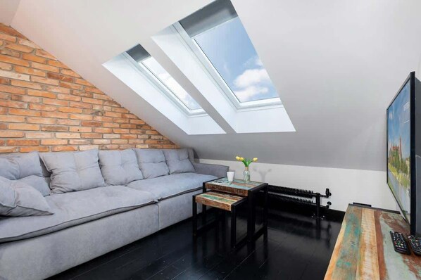 A cozy living room with a large gray sectional sofa positioned under a skylight. The exposed brick wall adds warmth, while the slanted ceiling gives it a unique loft-like feel.