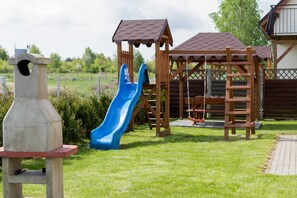 A well-equipped playground in a backyard, featuring a wooden slide and swing set surrounded by green grass.