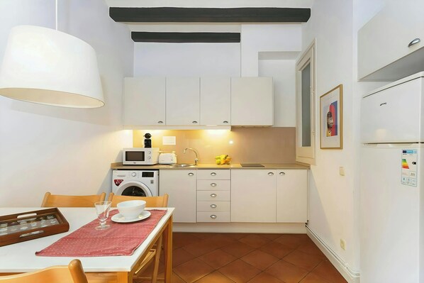 The apartment's kitchen features a modern design with white cabinets, a sleek countertop, and a tiled floor. The kitchen is fully equipped with a microwave, toaster, coffee maker, and a washing machine. It also has a refrigerator, a sink, and a stovetop. 