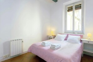 The bedroom features a queen-size bed with a pink and white striped duvet, providing a comfortable and inviting space for guests. The room is brightly lit with a large window overlooking a charming courtyard, offering a peaceful respite from the bustling 