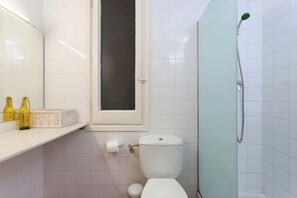The bathroom features white tiled walls and a frosted glass shower enclosure with a handheld showerhead.  The toilet is located next to the shower, with a window offering natural light. This bathroom is a clean and functional space for guests to refresh t