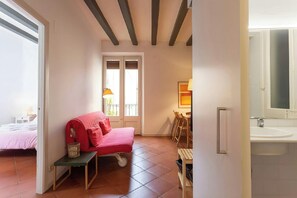 The living room features a red sofa bed with a side table and a glimpse into a bedroom. The room's exposed wooden beams and terracotta tile floors add a charming touch of character. A door leads to a bathroom, which is partially visible with a white sink 