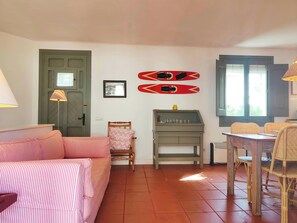 The living room features a comfortable pink and white striped sofa, a wooden armchair, and a rustic wooden dining table with wicker chairs, creating a warm and inviting ambiance. The tiled floor adds a touch of traditional charm. Two red water skis hang o