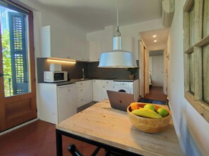 The apartment's dining table features a light wood top with a black metal frame. It's set for two. There is  a glimpse of the kitchen and a hallway in the background. The kitchen features white cabinets, a stainless steel sink, and a washing machine. The 